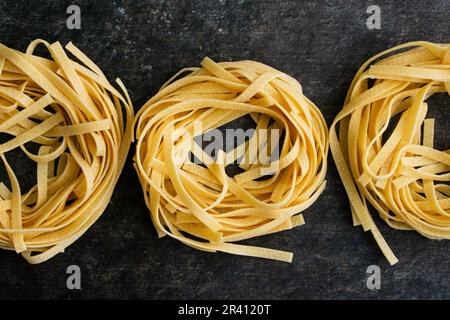 Eine Reihe von drei Tagliatelle Pasta-Nestern auf dunklem Hintergrund: Getrocknete Nudeln, die in einer Reihe von Nudelnestern angeordnet sind, die direkt von oben betrachtet werden Stockfoto