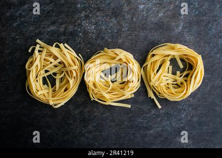 Eine Reihe von drei Tagliatelle Pasta-Nestern auf dunklem Hintergrund: Getrocknete Nudeln, die in einer Reihe von Nudelnestern angeordnet sind, die direkt von oben betrachtet werden Stockfoto