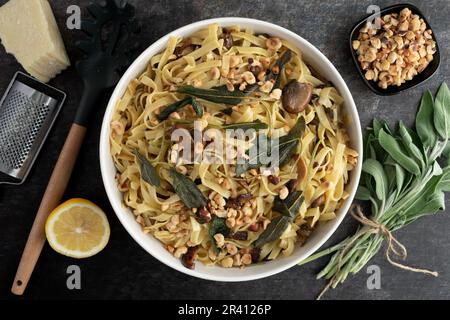Tagliatelle mit Pilzen, Salatbutter und gerösteten Haselnüssen in einer Servierschüssel: Eine große Schüssel mit Nudeln, umgeben von Zutaten und einem Nudellöffel Stockfoto