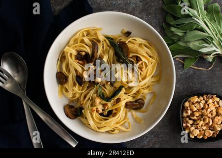 Tagliatelle mit Pilzen, Salbeibutter und gerösteten Haselnüssen in einer Pastaschale: Eine Portion Nudeln garniert mit gebratenen Salbeiblättern in einer flachen Schüssel Stockfoto