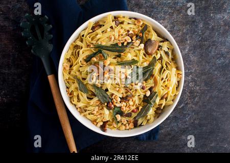 Tagliatelle mit Pilzen, Salatbutter und gerösteten Haselnüssen in einer Servierschüssel: Eine große Keramikschüssel mit garnierten Nudeln mit einem Nudellöffel Stockfoto