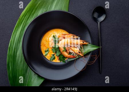 Meeresfrüchte-Cremesuppe mit Garnelen, die mit tropischen Blättern verziert sind. In schwarzer Platte, auf schwarzem Hintergrund. Draufsicht, Kopierbereich. Stockfoto