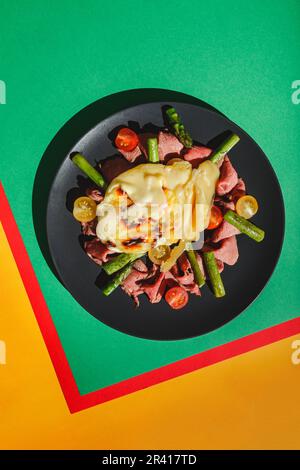 Köstlicher Raclette-Käse serviert über Rinderbraten mit Tomaten und Spargel, Gericht auf grünem Hintergrund. Hartes Licht, tiefer Schatten Stockfoto