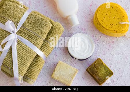 Grüne, saubere und trockene gefaltete Handtücher auf strukturierter Oberfläche mit Shampoo, Badeschwamm und natürlichen Olivenölseifen. Stockfoto