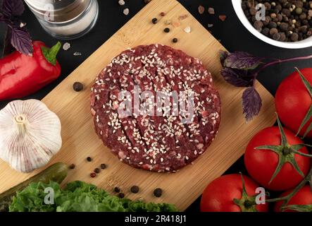 Rohe Hamburger-Pasteten mit Zutaten zum Kochen auf schwarzem Hintergrund Stockfoto