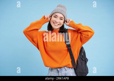 Ein süßes lächelndes asiatisches Mädchen zieht einen warmen Hut an, um nach draußen zu gehen, geht mit einem Rucksack im orangefarbenen Pullover spazieren, blauer Hintergrund Stockfoto