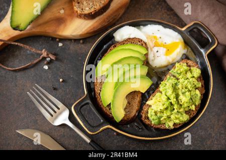 Vollkornbrot, Avocado in Scheiben und pochiertes Ei auf Steinhintergrund. Gesundes Frühstückskonzept, Sandwich mit Avocado und Stockfoto