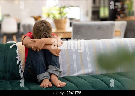 Ein trauriger, weißer Junge, der sich in den Knien umarmt, während er im Wohnzimmer auf dem Sofa sitzt, Kopierraum Stockfoto