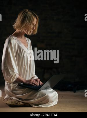 Frau und Laptop in einem leeren dunklen Raum. Deadline, viel Arbeit, Privatsphäre. Eine Frau mittleren Alters in einem weißen Hemd. Stockfoto