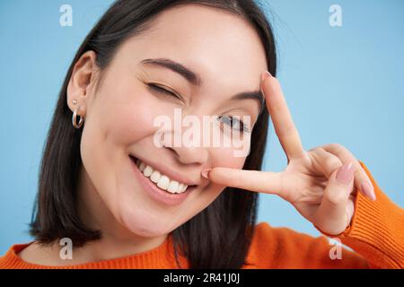 Nahaufnahme des Porträts einer positiven asiatischen Frau, zwinkert und zeigt Peave, V-Zeichen-Geste, klebt Zunge und Lächeln, steht über blauem Bac Stockfoto