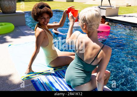 Fröhliche, vielfältige weibliche Freunde, die eine Poolparty feiern, einen Toast ausbringen und im Garten lächeln Stockfoto