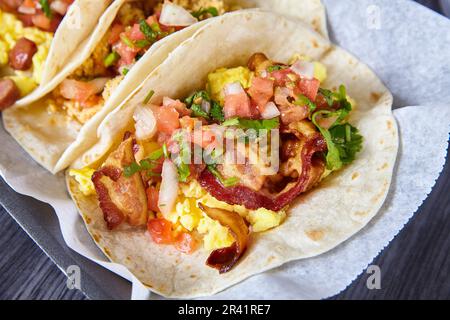 Mexikanisches Frühstück drei Tacos Wurst Eier Speck Tomaten Schnittlauch Mehl Tortillas Essen auf weißem Papier Stockfoto