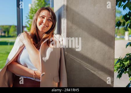 Glückliche Geschäftsfrau, die an der Wand in einem sonnigen Büro steht. Junger lächelnder Chef, entspannt. Frau trägt Anzug Stockfoto