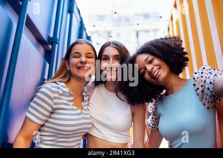 Drei süße Teenager-Mädchen in lässiger Kleidung auf bunten Versandbehältern. Stockfoto