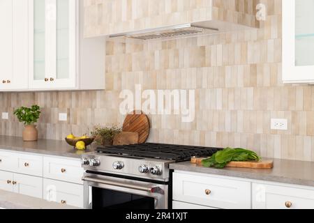 Ein Küchenofen und eine Kapuze mit braunen rechteckigen Fliesen, Edelstahlofen, weißen Schränken und gemütlichem Dekor auf Arbeitsflächen aus grauem Stein. Stockfoto