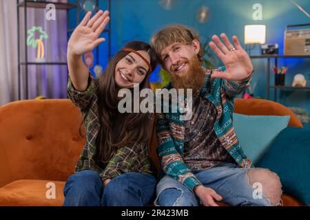 Glückliche junge Familienehe Hippie Paar Frau lächelt freundlich vor der Kamera und winkt Hände, hallo, hallo, grüßt oder auf Wiedersehen, willkommen zu Hause. Mann und Frau zusammen auf dem Sofa im Zimmer Stockfoto
