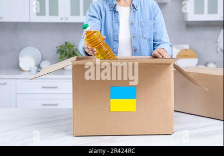 Humanitäre Hilfe für die Ukraine. Eine Frau, die eine Flasche Öl in eine Spendenbox am weißen Tisch im Haus legt, Nahaufnahme Stockfoto