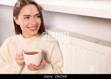 Frau hält Becher mit heißem Getränk in der Nähe des Heizkörpers in Innenräumen Stockfoto