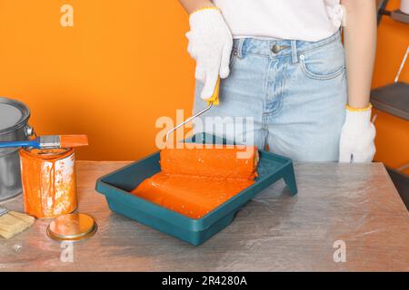 Frau nimmt orangefarbene Farbe mit Rolle vom Tablett am Tisch, Nahaufnahme Stockfoto