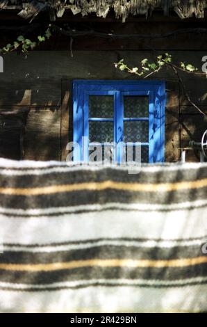 Maramures County, Rumänien, 2001. Einfaches Holzfenster auf einem lokalen Haus. Eine traditionelle Wolldecke, die am Zaun trocknet. Stockfoto