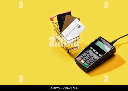 POS-Zahlungsterminal und Warenkorb mit Bankkarten auf gelbem Hintergrund. Minimales Finanzdienstleistungskonzept. Trendige Schatten, Stockfoto