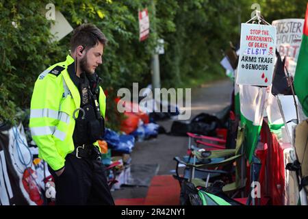 Ein Polizist liest Schilder im offiziellen Protestbereich gegenüber dem Haupttor der taktischen UAV-Systeme. Demonstranten von Palestine Action protestieren seit Mai 1 2023 außerhalb der UAV-Fabrik für taktische Systeme in Leicester. Sie sagen, sie bleiben hier, bis das taktische UAV-System abfährt. Die Polizei hat eine Sektion 14 erlassen, die es Demonstranten erlaubt, ein kleines offizielles Protestgebiet gegenüber der Fabrik zu benutzen. Jeder Protest außerhalb dieses Gebiets verstößt gegen den Befehl und wird zu einer Verhaftung führen. Demonstranten erheben Einspruch gegen Waffen, die im Vereinigten Königreich von der israelischen Verteidigungsgesellschaft Elbit Systems und ihrem Subventionar hergestellt werden Stockfoto