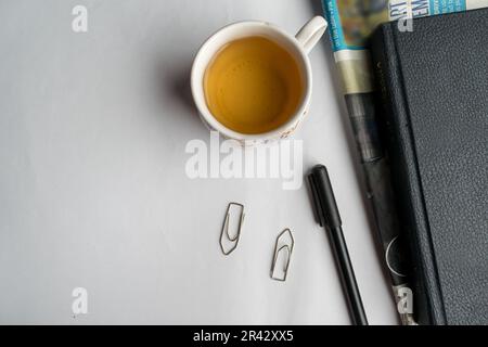 Flacher Schreibtisch mit Teetasse, Notizbuch, Stift, Zeitung und Büroklammern auf weißem Hintergrund. Stockfoto