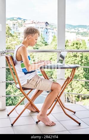Ein junger Junge sitzt auf dem Hotelbalkon und surft auf einem grauen Laptop im Internet. Der Schuljunge verbringt gern Sommerferien und spielt gern Spiele Stockfoto
