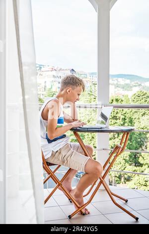 Ein junger Junge sitzt auf dem Hotelbalkon und surft auf einem grauen Laptop im Internet. Der Schuljunge verbringt gern Sommerferien und spielt gern Spiele Stockfoto