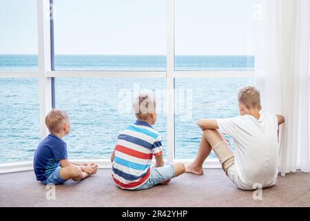 Jungen Geschwister genießen es, das azurblaue Meer vom Panoramafenster im Hotelzimmer aus zu beobachten. Blonde Brüder verbringen Sommerferien zusammen am Meer Stockfoto