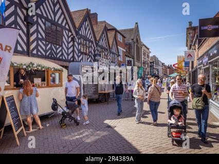 Gäste genießen die Sonne, während sie entlang einer belebten Straße mit Geschäften spazieren und während des Lichfield Food Festivals Speisen und Getränke an den Verkaufsständen aufstellen. Stockfoto