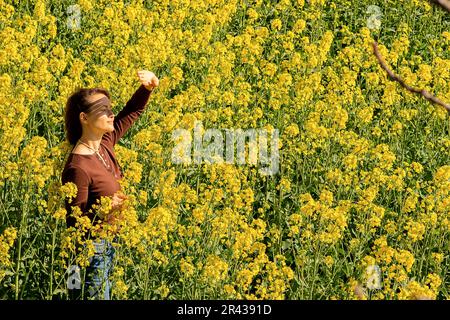 Eine junge Weiße geht durch ein blühendes, gelbes Rapsfeld, und wenn die Sonne hell scheint, blickt sie durch ihre Handfläche in die Ferne. Stockfoto