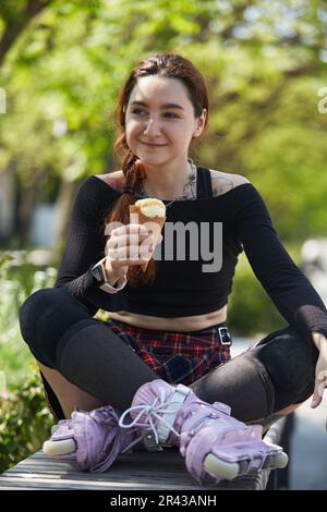 Fröhliche Skateboarderin, die ein Eis auf einer Bank in einem grünen Park isst. Glücklicher junger erwachsener Rollschuh, der moderne Inline-Skates trägt und leckt Stockfoto
