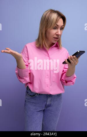 Eine junge blonde Frau mit schulterlangem Haar in einem modischen rosa Hemd liest Nachrichten vom Telefon Stockfoto