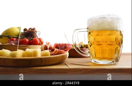 Bierbecher und Vorspeise mit Schinken und Käse, Oliven, Tomaten und Erdnüsse, zubereitet auf einem Holztisch und mit einem weißen, isolierten Hintergrund. Vorderansicht. Stockfoto