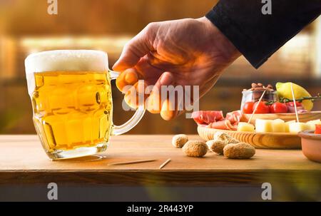 Mann mit einer Tasse Bier in der Hand auf einer Holzbank mit Vorspeise, zubereitet in einer rustikalen Küche. Vorderansicht. Stockfoto