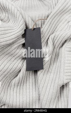 Leerer schwarzer Pappanhänger mit Schnur auf grauem Pullover. Beschriften Sie eine Kleidung mit einem leeren Modell, um Ihr Design zu platzieren. Preisschild, Verkauf, Adressetikett. Einkaufen Stockfoto