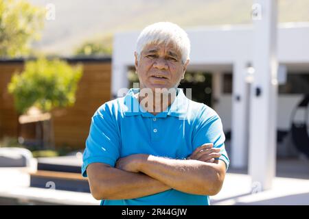 Porträt eines glücklichen älteren, birassischen Mannes mit weißen Haaren und gekreuzten Armen im sonnigen Garten Stockfoto