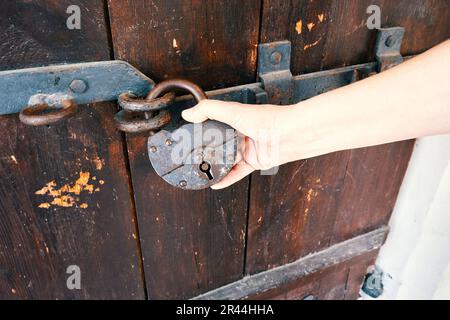 Alte Holztür mit rostigem Schloss. Nahaufnahme einer weiblichen Hand, die ein Vorhängeschloss hält Stockfoto