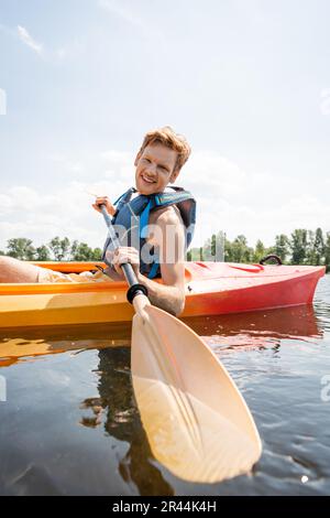 Ein fröhlicher rothaariger Mann in der Schwimmweste hält ein Paddel und schaut in die Kamera, während er Zeit auf dem See mit dem Kajak während der Wassersportangebote auf verbringt Stockfoto