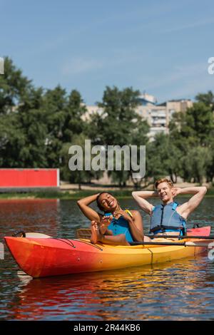 Frohe und barfuß afroamerikanische Frau und lächelnder rothaariger Mann in Schwimmwesten, der sich während des Sommerwochenendes mit geschlossenen Augen im sportlichen Kajak entspannt Stockfoto