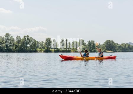 Seitenansicht einer afroamerikanischen Frau mit einem jungen Mann in Schwimmwesten, die im Sommer im sportlichen Kajak am grünen Flussufer entlang paddelt Stockfoto