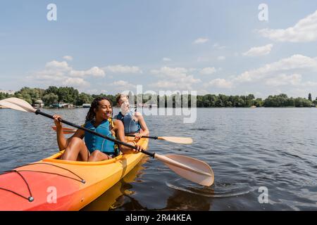 Sorglose und charmante afroamerikanische Frau und junger rothaariger Mann in Schwimmwesten segeln in sportlichem Kajak auf ruhiger Wasseroberfläche unter blauem Himmel mit C. Stockfoto