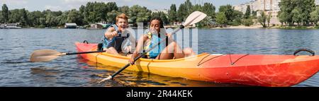 Ein überfreulicher rothaariger Mann zeigt mit dem Finger auf eine afroamerikanische Frau in einer Schwimmweste, die ein Paddel hält, während sie im Kajak am grünen Flussufer entlang segelt Stockfoto
