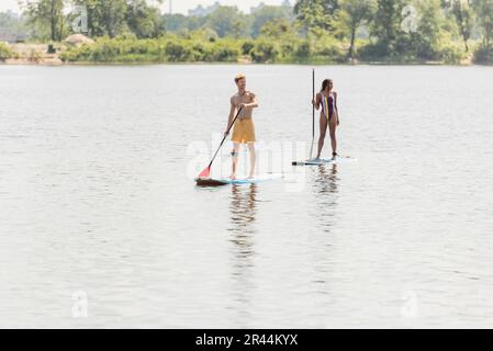 Ein junger Sportler und eine afroamerikanische Frau im gestreiften Badeanzug, die im Sommer auf Sup Boards mit Paddeln auf dem See segeln Stockfoto