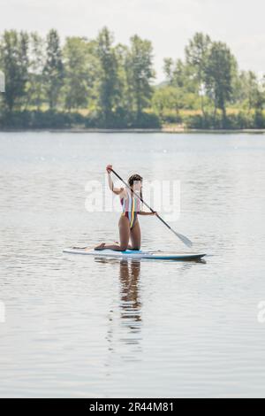 Eine lange, aktive und sportliche afroamerikanische Frau in farbenfrohen Badeanzügen balanciert auf den Knien, während sie am Sommertag auf dem Sup-Board segelt und paddelt Stockfoto