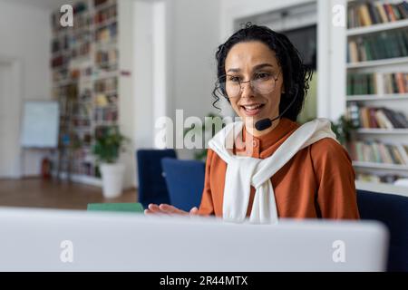 Lehrer führt Unterricht online durch, Frau mit Headset für Videogespräche lächelt und erklärt Vorlesungen online mit Laptop für Fernkommunikation und Lernen, sitzt am Schreibtisch in der Universitätsbibliothek am Schreibtisch. Stockfoto