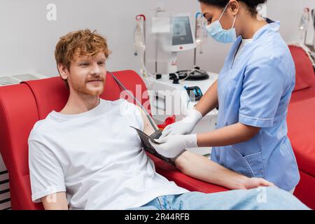 Multirassische Krankenschwester in medizinischer Maske und Latexhandschuhen justieren Blutdruckmanschette am Arm eines Rotkopfspenders, der in der Nähe des Transfusionsgeräts in der Klinik sitzt Stockfoto