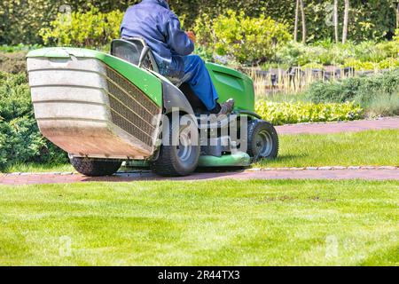 Ein Mitarbeiter eines öffentlichen Versorgungsunternehmens in blauem Overall mäht das grüne Gras des Rasenmähers. Verschwommener Hintergrund eines Frühlingsgartens. Speicherplatz kopieren. Stockfoto