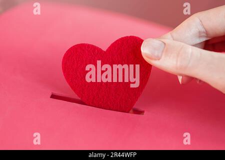 Die weibliche Hand steckt Red Heart in die rote Spendenbox. Konzept der aufrichtigen Hingabe zum Glauben. Konzept der Spende, Lebensrettung oder Wohltätigkeit. Nahaufnahme Stockfoto